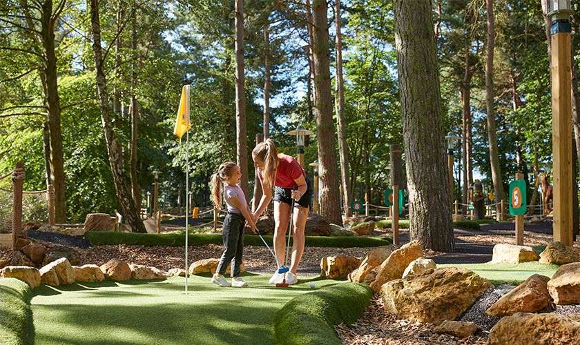 Two people play mini-golf, one teaching the other to putt, on a green with a flagstick, surrounded by rocks and trees; course signs display “5.”