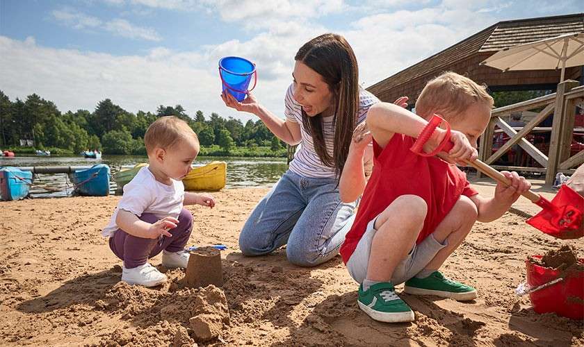 Children build sandcastles, scooping sand with red shovels, while a woman holds a blue bucket and watches. Context: sandy lakeside beach with kayaks, wooden pavilion, trees, and partly cloudy sky.