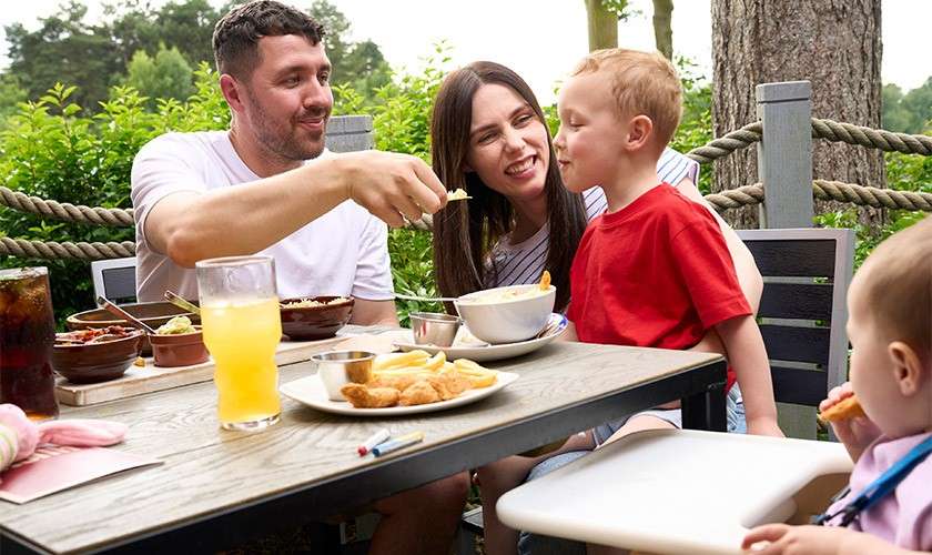 Family dining; a man feeds a spoonful to a young boy as a woman smiles. A toddler sits in a high chair. Plates and drinks rest on a patio table.