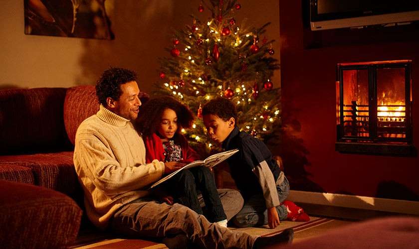Man reads an open book to two children, seated on a rug. Warm living room context: lit Christmas tree, glowing fireplace, sofa and soft lighting creating a cozy holiday atmosphere.