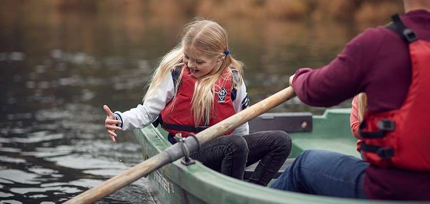 Girl in a red life jacket touches the water as another person rows, in a small green rowboat on a calm lake with blurred autumn foliage in the background.