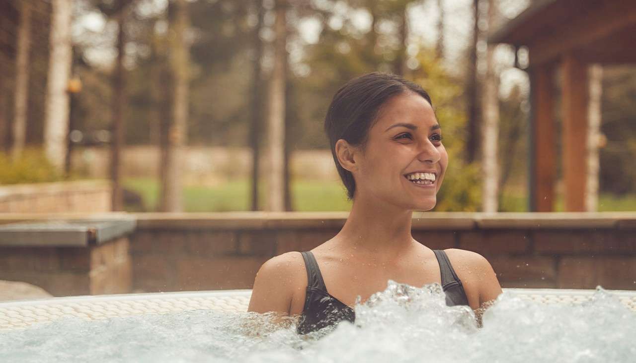 A woman smiles while sitting in a bubbling hot tub outdoors, shoulders above water, wearing a black swimsuit; trees, patio walls, and a wooden structure appear blurred in the background.