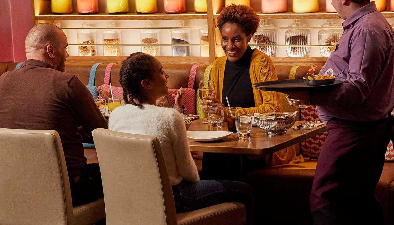 Diners converse and smile while a waiter serves food at a wooden table; glasses and plates set; cozy restaurant with patterned booths and shelves of jars and glowing colored containers.