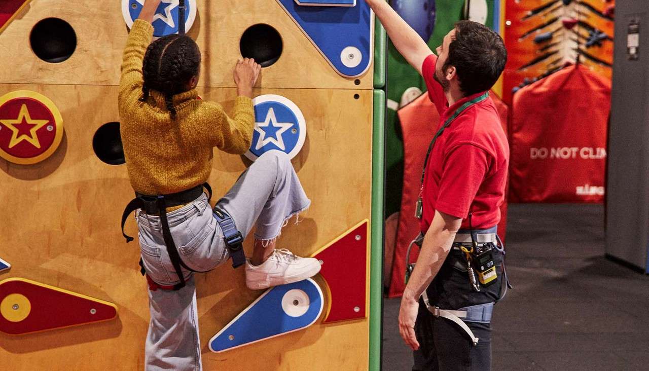 Climber ascends a colorful indoor wall, gripping round star holds while wearing a harness. Instructor beside points upward, guiding. Padded gym and safety gear surround. Visible text: DO NOT CLIMB.
