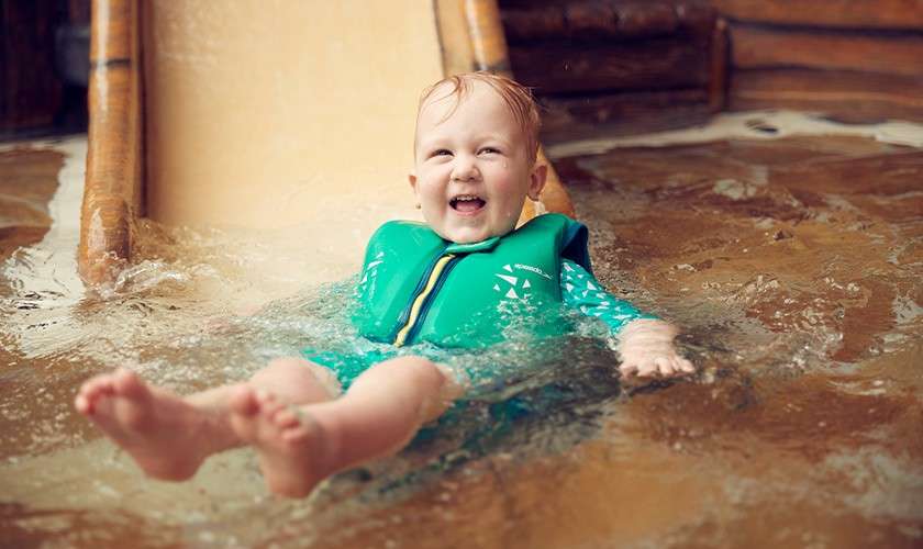 Smiling toddler wearing a green life vest leans back and splashes into shallow water after descending a small wooden slide inside an indoor water park.