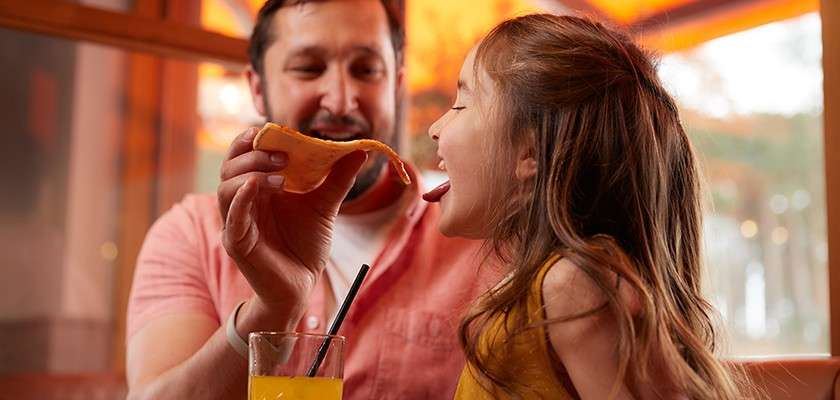 Man holds a cheesy pizza slice, feeding a smiling girl as she opens her mouth; a glass of orange drink with a straw sits upfront in a warmly lit restaurant.