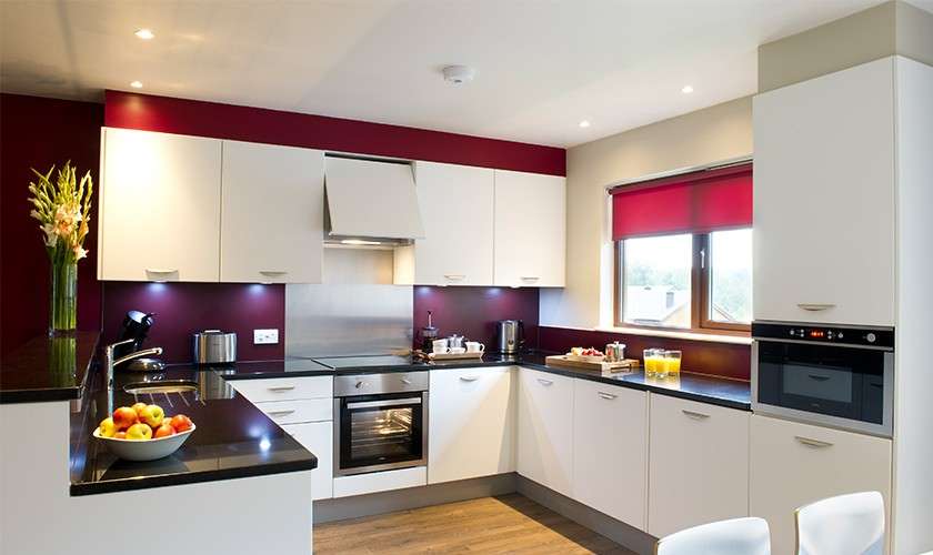 Modern kitchen—white cabinets frame a stainless oven and stovetop—sits under recessed lights, with black countertops, red backsplash and window blind, sink, appliances, fruit bowl, and glasses of juice on counters.