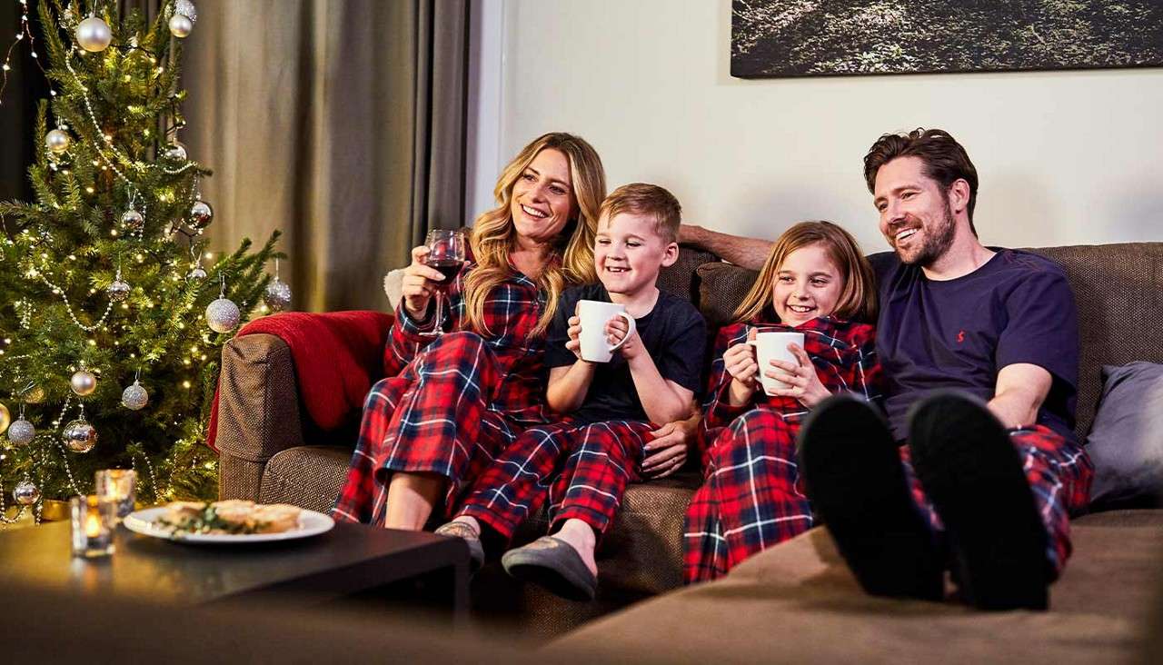 Family on a sofa relaxes, drinking from mugs and a wineglass, smiling. They wear matching red plaid pajamas in a living room with lit Christmas tree, candles, blanket, and artwork.