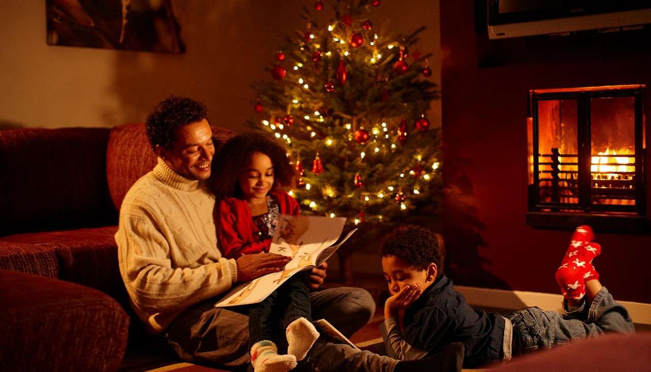 Father and two children read a picture book on the floor. One child lounges on a rug. Nearby, a fireplace glows beside a decorated Christmas tree in a warm room.