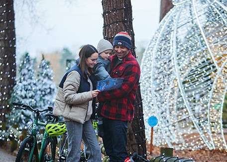 Family huddles while examining a booklet; the child held between adults. They stand among bicycles and helmets in a forest decorated with holiday lights and a glowing spherical structure.
