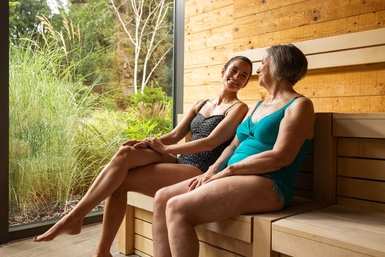 Two swimsuit-clad women sit and chat on wooden benches inside a sauna-like spa room beside a large window overlooking lush greenery.