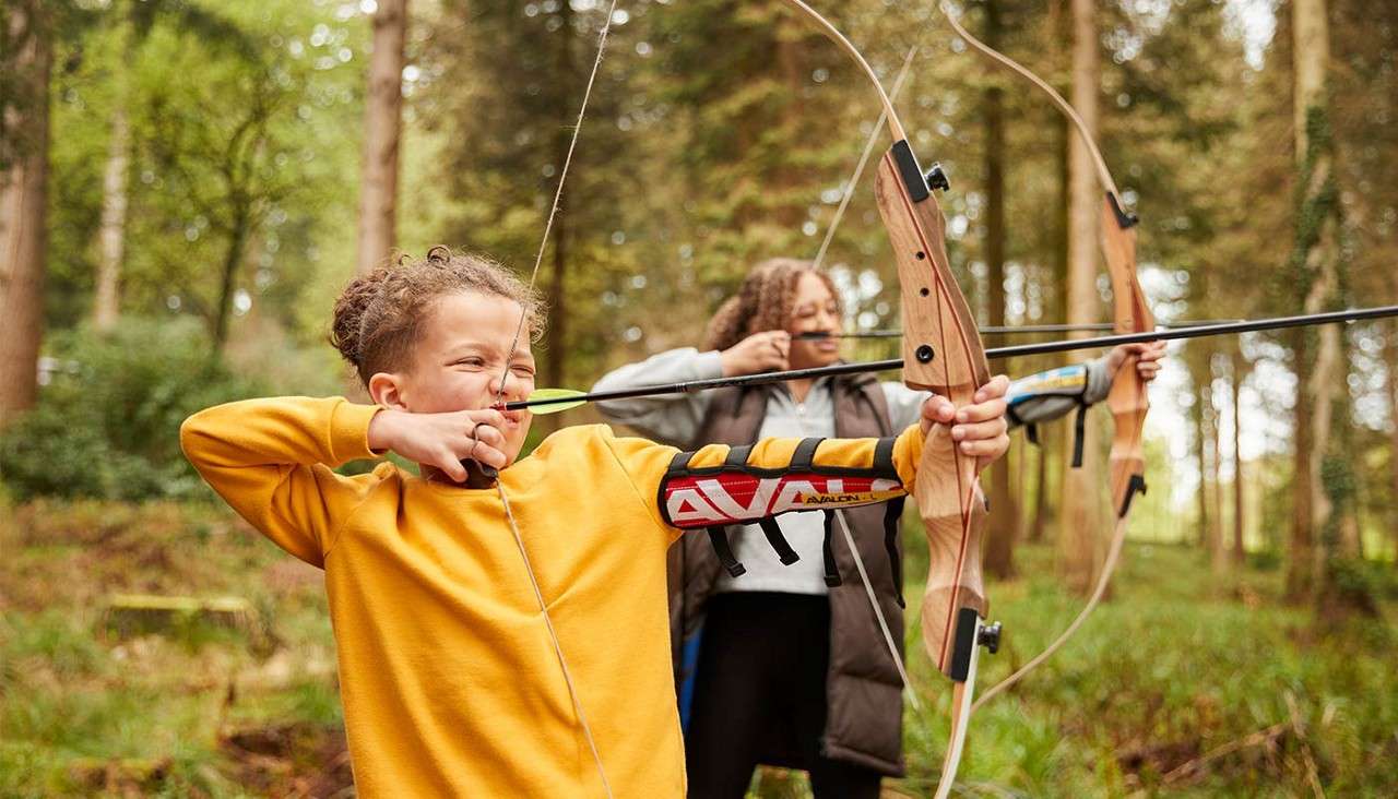 Children archers pull back bowstrings, aiming wooden recurve bows. In a sunlit forest, they concentrate during an outdoor lesson; the front child's armguard shows the text: "AVALON".