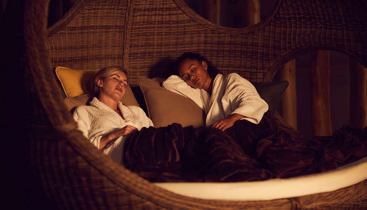 Two people in white bathrobes rest with eyes closed, nestled against pillows under dark blankets, inside a large wicker pod lounge, in warm, dim spa-like surroundings with wooden posts.