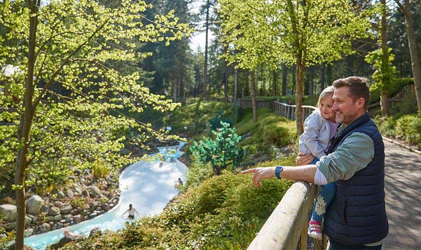 Father holding child leans on wooden railing, pointing toward a winding lazy river where people float on tubes, surrounded by lush green trees and a sunlit forest path.
