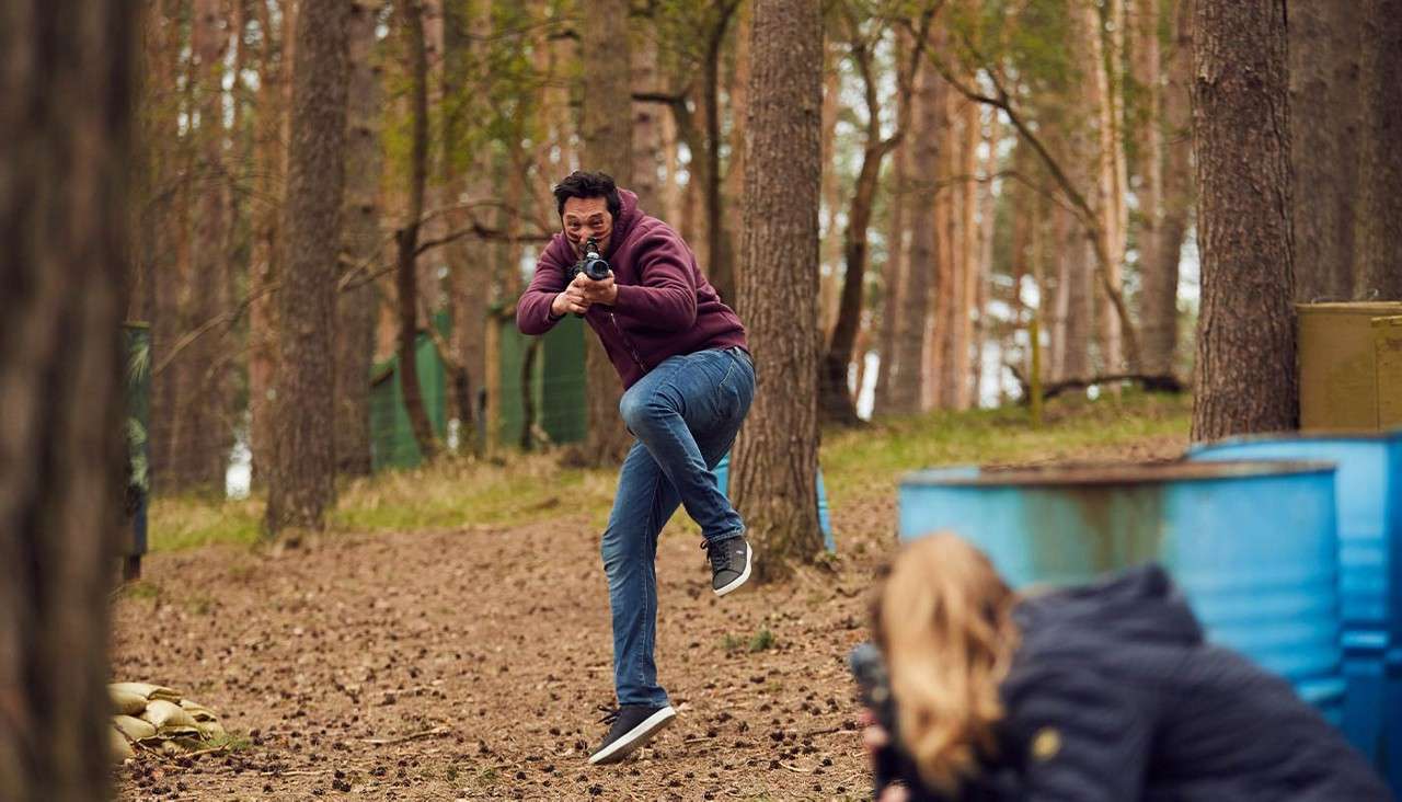 Man aims a paintball gun while running; another player crouches behind blue barrels; forest setting with tall trees, leafy ground, and green netting obstacles.