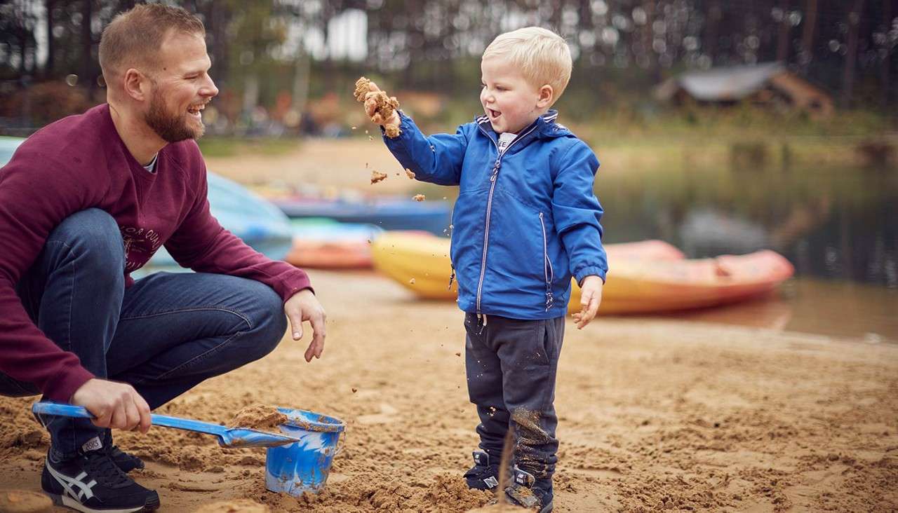 Child drops wet sand; nearby adult crouches shoveling. Context: sandy lakeshore with blue bucket and shovel, colorful kayaks on shore, calm water and forested background.
