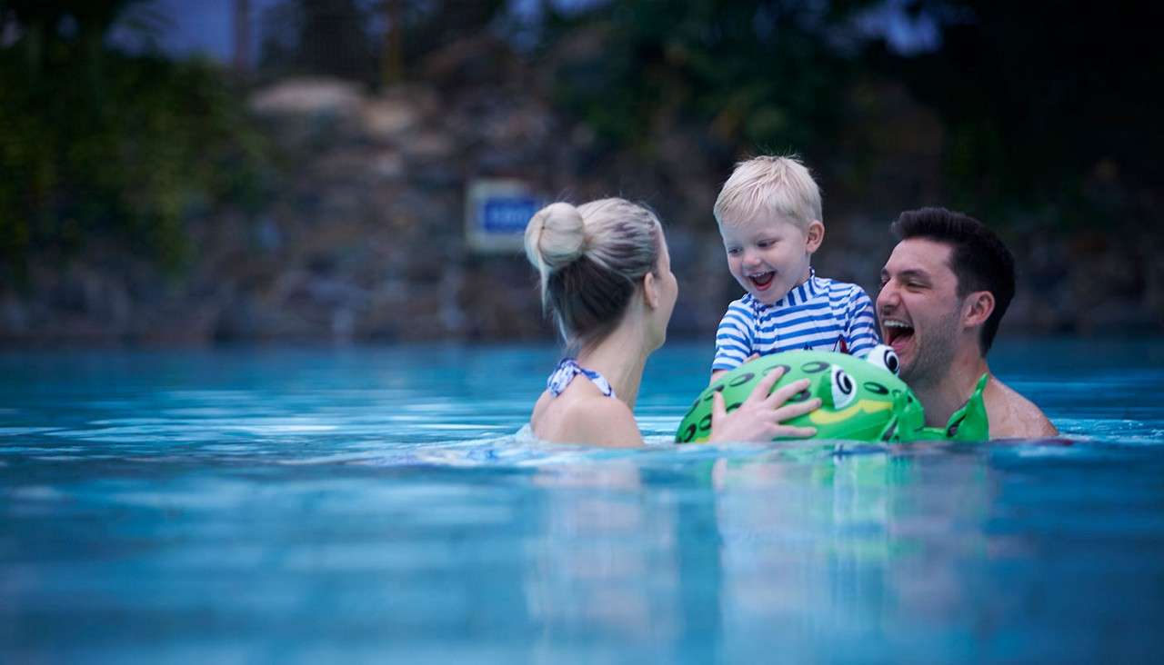 A laughing child rides a green turtle float while two adults hold him, swimming together in a large outdoor pool at dusk, with trees and rocks in the background.