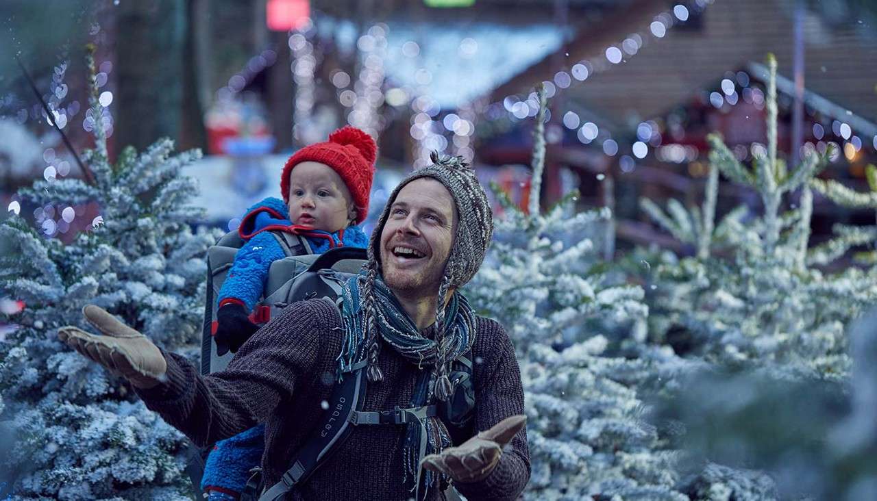 Adult wearing knit hat carries toddler in a backpack, smiling and catching falling snow. Context: snow-covered evergreen trees and festive string lights line a wintry outdoor market or tree lot.