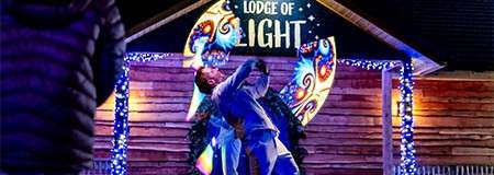 Two performers dance, one lifting the other, in front of a wooden building decorated with colorful lights and a sign reading LODGE OF LIGHT at night, while spectators watch.