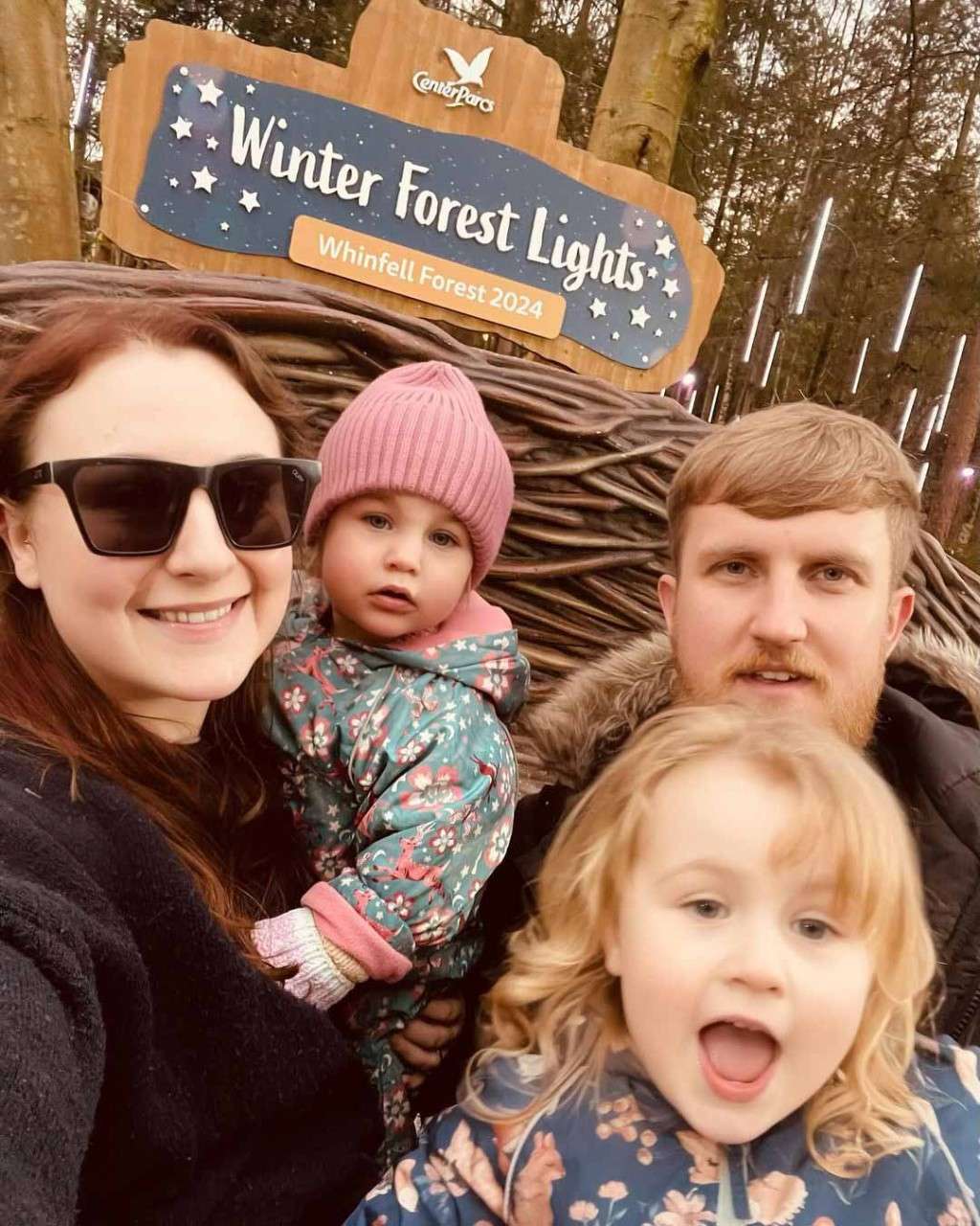 A family taking a selfie in front of the winter forest lights sign.