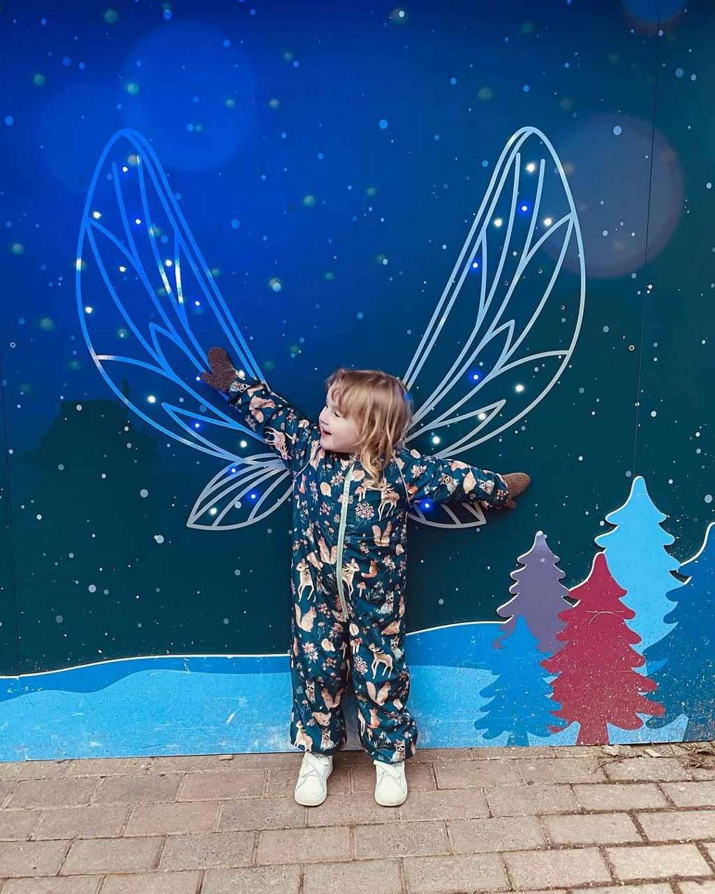 Child stands arms outstretched, aligning with glowing illustrated wings. Starry blue mural backdrop features snow, stylized pine trees, and dots; child wears patterned snowsuit, mittens, and shoes on brick pavement.