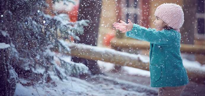 Child extends outstretched hands, catching falling snowflakes. Set in a snowy outdoor scene beside evergreen trees and a wooden fence, wearing a teal coat and pink knit hat, cheeks rosy.
