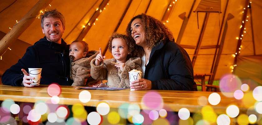 A family of four sits at a wooden counter, sipping hot drinks and smiling, inside a warmly lit tent decorated with string lights; colorful bokeh dots blur the foreground.