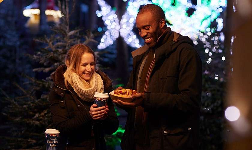 Two people wearing winter coats share hot drinks and a plate of food, smiling under night-time holiday lights among trees. Text on cups: "Winter ... Lights" (partially unreadable).
