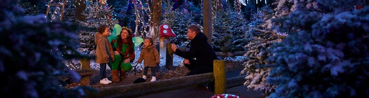 Family with two children and a costumed fairy smile and chat among snow-dusted Christmas trees and lights, by a path, while one adult kneels and gestures beside red-and-white mushroom decorations.