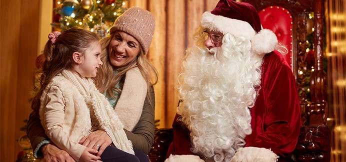 Child sits on a woman's lap, smiling and talking with a man dressed as Santa. They face each other beside a Christmas tree, near an ornate red chair.