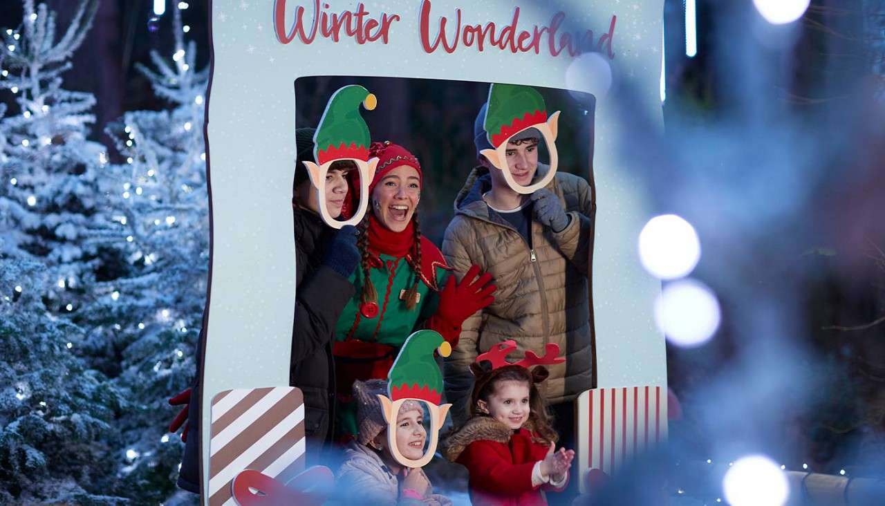 Group of children and adults pose and laugh through elf-face cutouts in a festive photo board labeled "Winter Wonderland," surrounded by snowy Christmas trees and string lights at night.