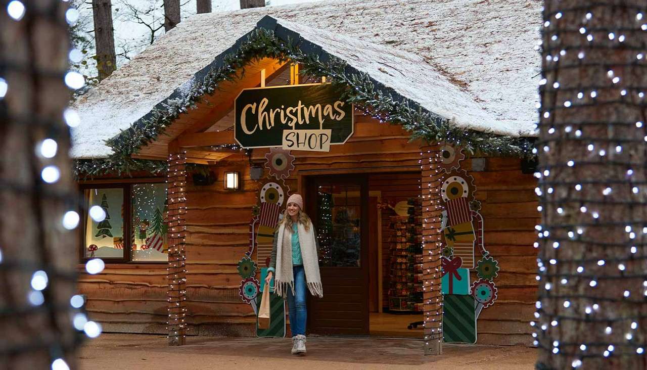 A bundled shopper walks out holding a paper bag, beneath a sign reading "Christmas SHOP"; context: snow-covered log cabin decorated with garlands and string lights, surrounded by trees.