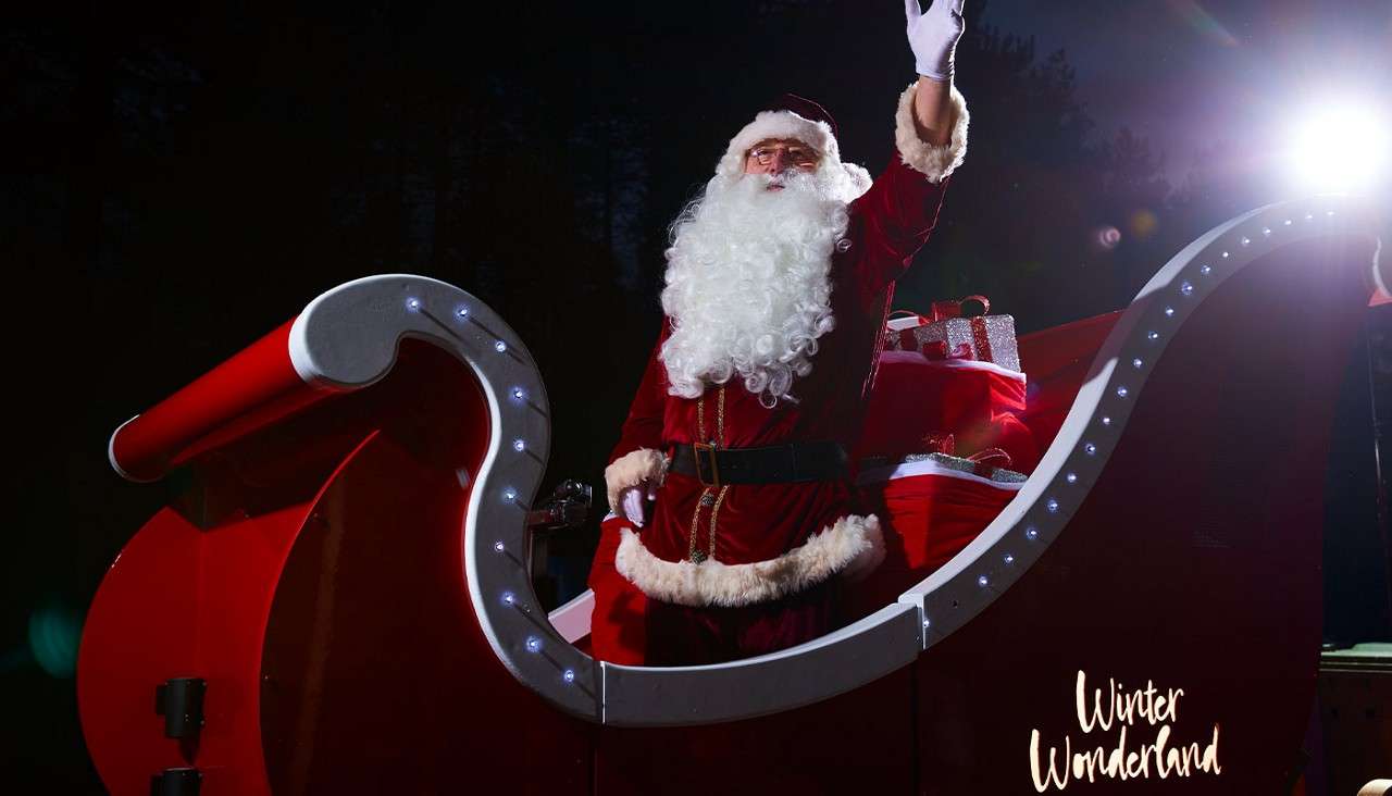 Santa Claus waves from a red, LED-lit sleigh, gifts stacked behind him, under a bright spotlight at night with dark trees. Text: Winter Wonderland.