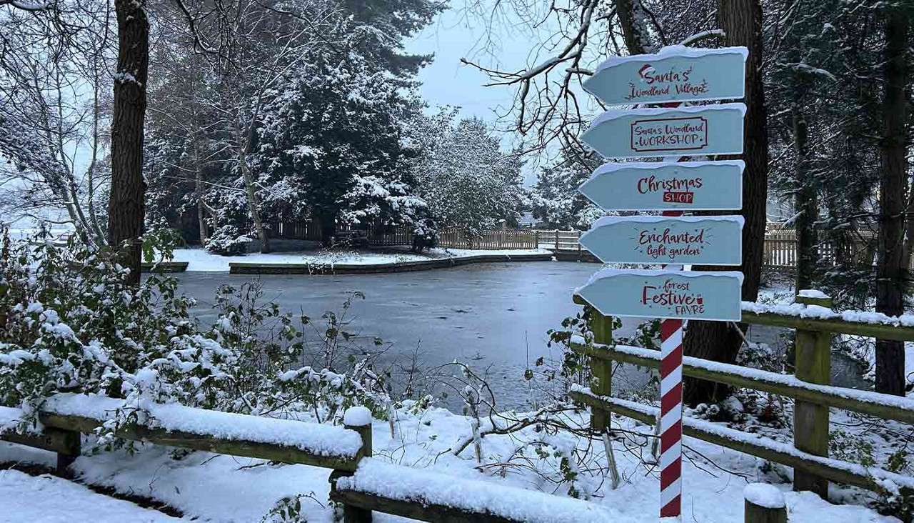 Striped signpost points directions in a snowy forest clearing near a frozen path and fence. Text: "Santa's Woodland Village," "Santa's Woodland Workshop," "Christmas Shop," "Enchanted light garden," "Forest Festive Fayre."