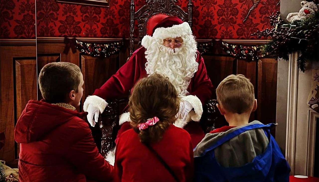 Santa Claus sits on an ornate chair, talking to three children facing him. The room is richly decorated for Christmas with red wallpaper, wooden paneling, garlands, and festive greenery.