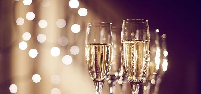 Champagne flutes hold bubbling sparkling wine, standing together on a table; in the background, warm bokeh lights glow softly, suggesting a festive indoor celebration.