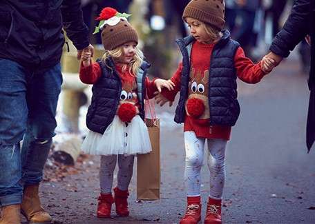 Two small children walk while holding adults’ hands, one carrying a paper bag, both wearing matching reindeer sweaters, pom‑pom hats, vests, leggings, and boots on a winter street.