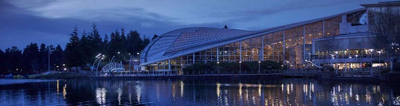 Glass-domed building glows with interior lights, reflecting on calm water; lampposts and a small arched structure line the shore; dusk sky and surrounding trees frame the modern waterfront complex.
