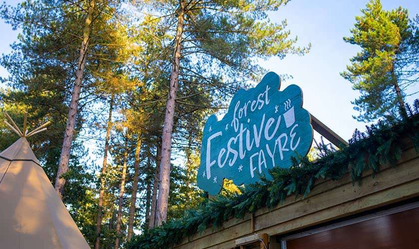 Signboard displays “forest Festive FAYRE,” mounted atop a wooden stall garlanded with evergreen boughs; teepee tent at left; tall pine trees and blue sky frame an outdoor forest market setting.