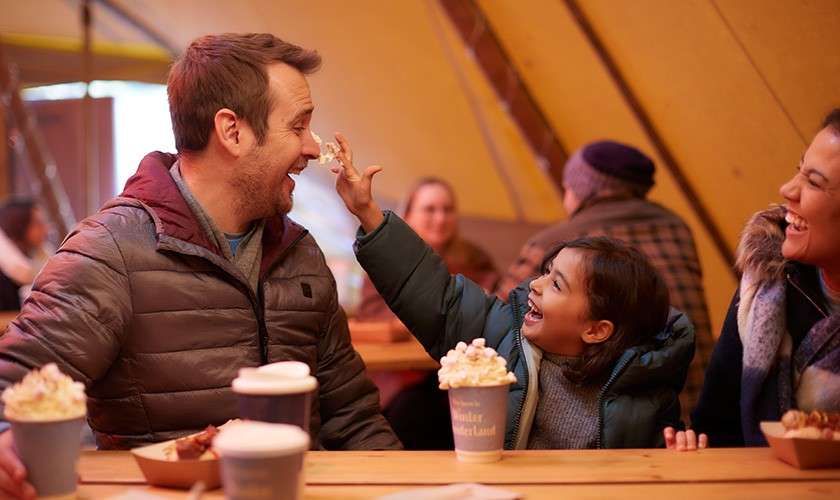 Child smears whipped cream on man's nose while laughing; adults smile nearby. Indoors at a warm cafe tent with hot chocolate and pastries on a wooden table. Text: “Winter Wonderland.”
