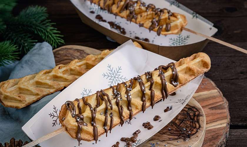Skewered waffle drizzled with chocolate rests in a paper tray; additional waffles on sticks sit nearby on wooden boards, with napkin, pinecones, and evergreen sprigs on a rustic wooden table.
