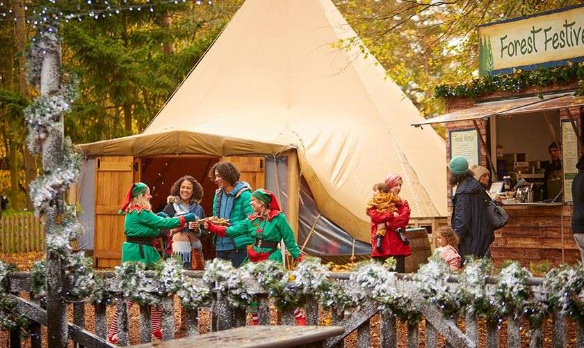 A family and group of elves standing outside of the Forest Fayre tepee with another family getting drinks from a stall