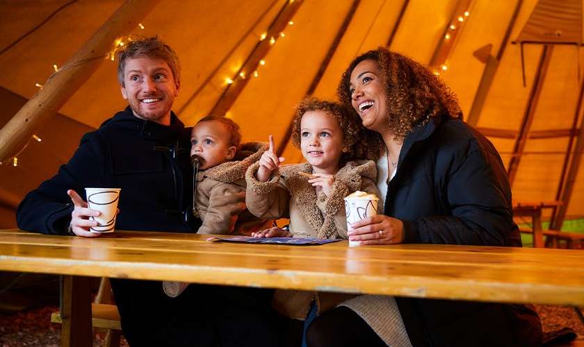 Family of four sits smiling and drinking from paper cups at a wooden table, child pointing. Context: cozy tent interior with warm lighting and string lights.