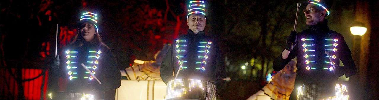 Three drummers wearing LED-lit vests and hats play illuminated drums, raising sticks midbeat, in a dark outdoor setting with trees, red and yellow lighting, and faint onlookers in the background.