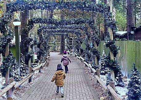 Two children run ahead under garland-wrapped wooden arches, along a brick path. Artificial snow dusts small Christmas trees and fencing on both sides, within a wooded park setting.
