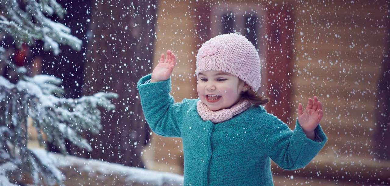 Child in teal coat and pink knit hat raises hands, smiling, catching falling snowflakes; outdoors amid snowfall, with a snow-dusted evergreen and blurred wooden building in the background.