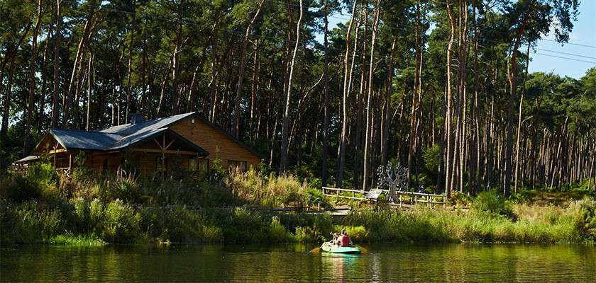 Two people paddle a small inflatable boat across calm water; behind them, a wooden cabin sits on a grassy lakeshore, framed by tall pine trees and a simple dock.
