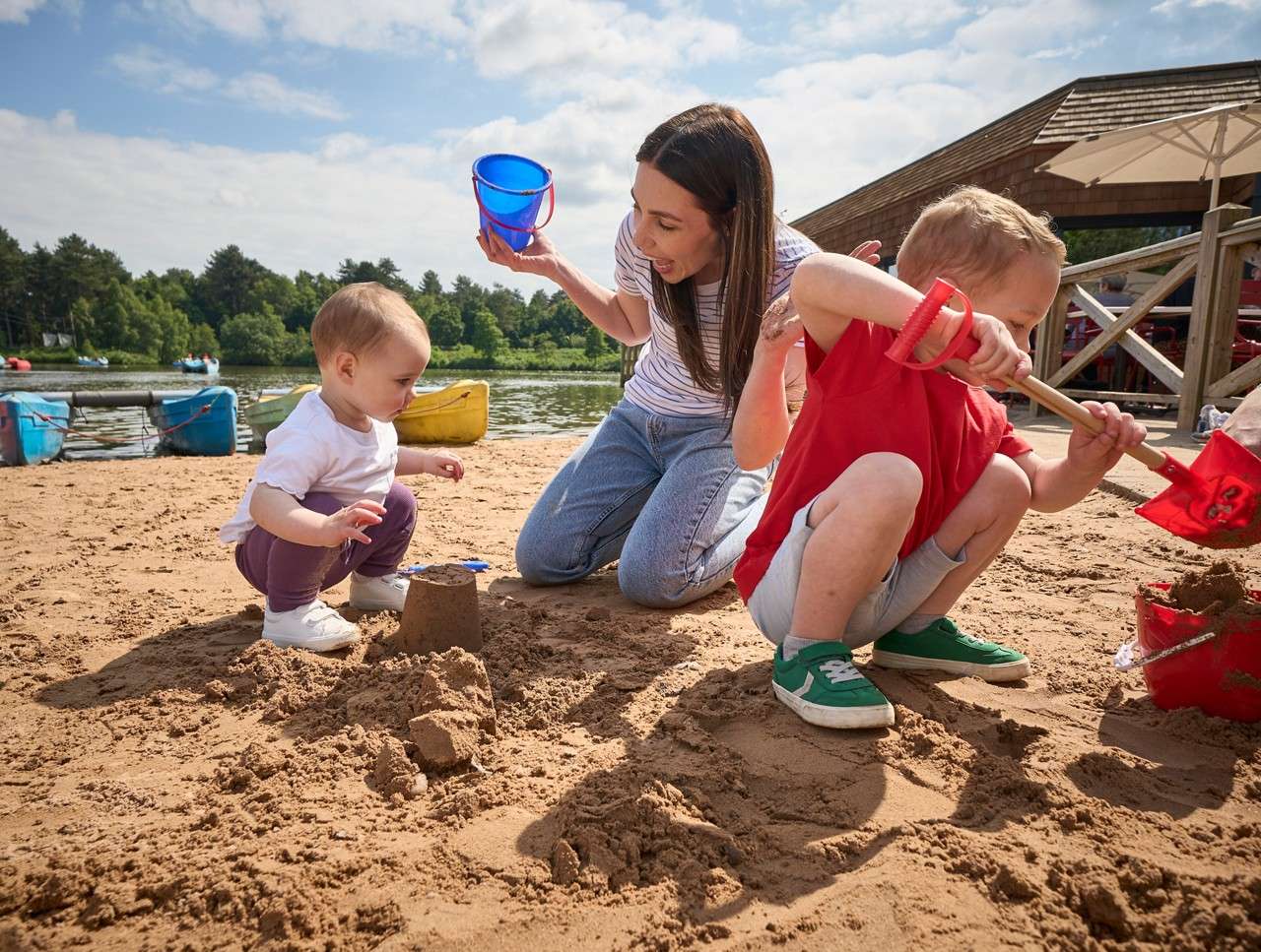 Children build sandcastles while an adult assists on a beach; one child digs with a red shovel, the other examines a small sand tower; lake, boats, and a wooden building behind.