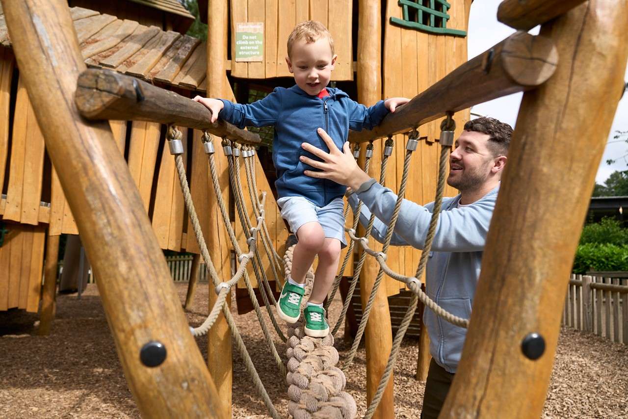 Child walks across a rope bridge while an adult steadies him; wooden playground structure surrounds with mulch ground. Sign reads: Do not climb on the external structure.