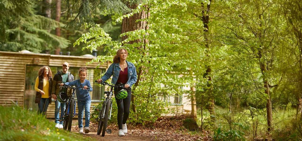 Family with bicycles pushes them along a dirt path, smiling, amid tall trees; a wooden cabin sits behind them in a sunlit, green forest.